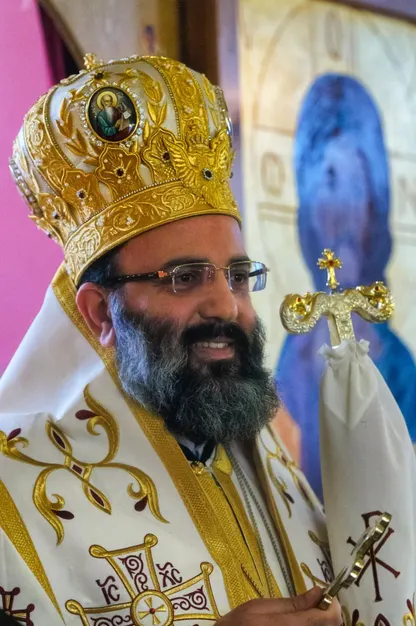 Metropolitan Silouan, smiling, in a gold mitre and white and gold vestments, before an icon